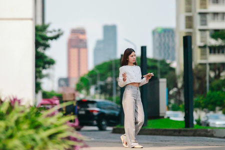 Asian woman dressed in modern business casual attire is seen writing in notebook while standing outdoors in urban setting. Conveying sense of focus, productivity, and creativity in city environmentの写真素材