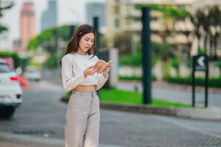 Asian woman dressed in modern business casual attire is seen writing in notebook while standing outdoors in urban setting. Conveying sense of focus, productivity, and creativity in city environmentの写真素材