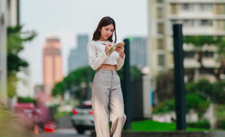 Asian woman dressed in modern business casual attire is seen writing in notebook while standing outdoors in urban setting. Conveying sense of focus, productivity, and creativity in city environmentの写真素材