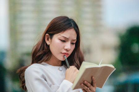 Asian woman dressed in modern business casual attire is seen writing in notebook while standing outdoors in urban setting. Conveying sense of focus, productivity, and creativity in city environmentの写真素材