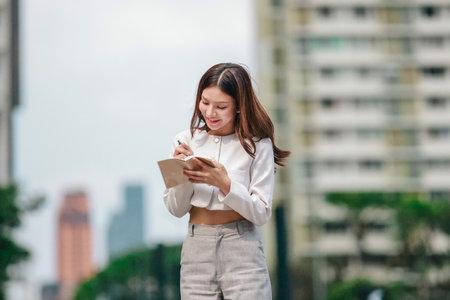 Asian woman dressed in modern business casual attire is seen writing in notebook while standing outdoors in urban setting. Conveying sense of focus, productivity, and creativity in city environmentの写真素材