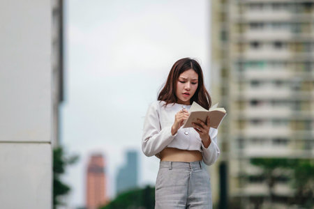 Asian woman dressed in modern business casual attire is seen writing in notebook while standing outdoors in urban setting. Conveying sense of focus, productivity, and creativity in city environmentの写真素材