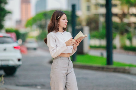 Asian woman dressed in modern business casual attire is seen writing in notebook while standing outdoors in urban setting. Conveying sense of focus, productivity, and creativity in city environmentの写真素材