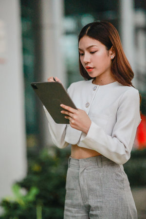 Asian woman in professional attire is standing outdoors, focused on using digital tablet with stylus. Capturing a modern business environment, highlighting technology and productivity in urban settingの写真素材