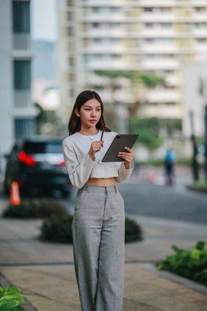 Asian woman in professional attire is standing outdoors, focused on using digital tablet with stylus. Capturing a modern business environment, highlighting technology and productivity in urban settingの写真素材
