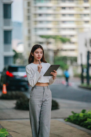 Asian woman in professional attire is standing outdoors, focused on using digital tablet with stylus. Capturing a modern business environment, highlighting technology and productivity in urban settingの写真素材