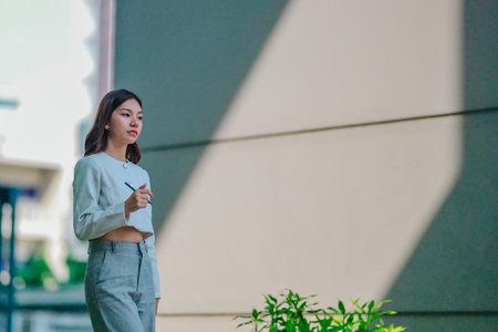 Asian woman in professional attire is standing outdoors, focused on using digital tablet with stylus. Capturing a modern business environment, highlighting technology and productivity in urban settingの写真素材