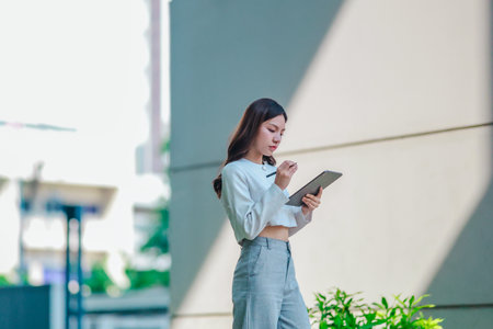 Asian woman in professional attire is standing outdoors, focused on using digital tablet with stylus. Capturing a modern business environment, highlighting technology and productivity in urban settingの写真素材