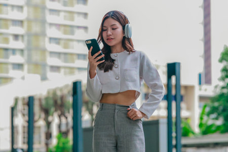 Young Asian woman wearing wireless headphones outdoors while looking at her smartphone. Surrounded...