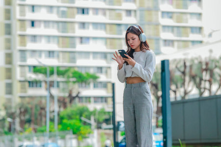 Young Asian woman wearing wireless headphones outdoors while looking at her smartphone. Surrounded by greenery and city buildings. Conveying modern, connected lifestyle blending technology and natureの写真素材