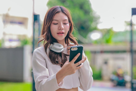 Young Asian woman wearing wireless headphones outdoors while looking at her smartphone. Surrounded...