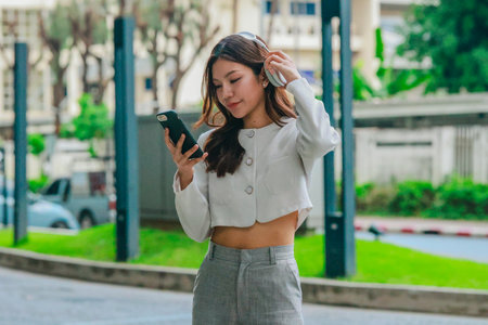 Young Asian woman wearing wireless headphones outdoors while looking at her smartphone. Surrounded by greenery and city buildings. Conveying modern, connected lifestyle blending technology and natureの写真素材