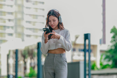 Young Asian woman wearing wireless headphones outdoors while looking at her smartphone. Surrounded...