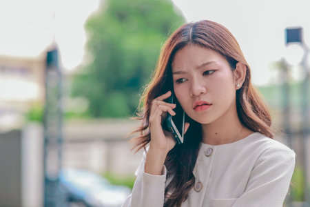 Asian woman appears frustrated while speaking on smartphone in outdoor urban environment. Her body...
