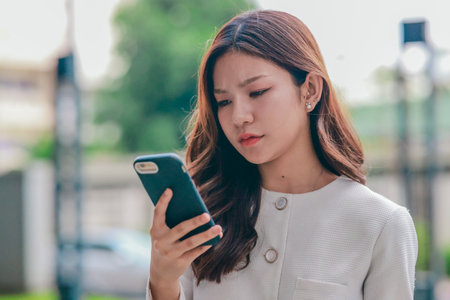 Asian woman appears frustrated while speaking on smartphone in outdoor urban environment. Her body...