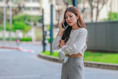 Asian woman appears frustrated while speaking on smartphone in outdoor urban environment. Her body...
