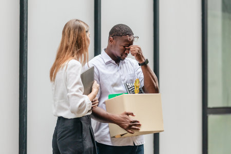 African male employee carries box of office belongings while female coworker encourages him outdoors. Reflecting job loss during an economic downturn and power of workplace support and resilienceの写真素材