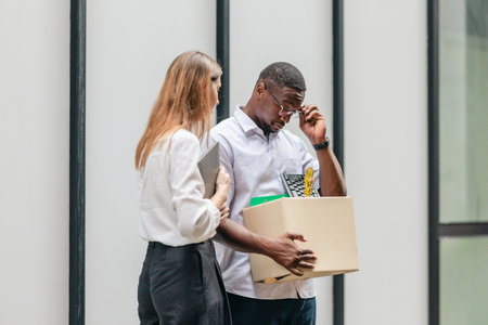 African male employee carries box of office belongings while female coworker encourages him outdoors. Reflecting job loss during an economic downturn and power of workplace support and resilienceの写真素材