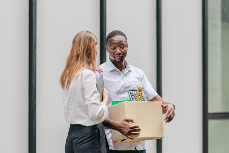 African male employee carries box of office belongings while female coworker encourages him outdoors. Reflecting job loss during an economic downturn and power of workplace support and resilienceの写真素材