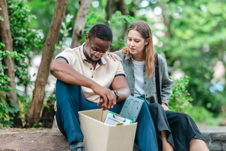 African male employee carries box of office belongings while female coworker encourages him...