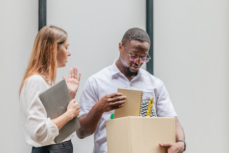 African male employee carries box of office belongings while female coworker encourages him outdoors. Reflecting job loss during an economic downturn and power of workplace support and resilienceの写真素材