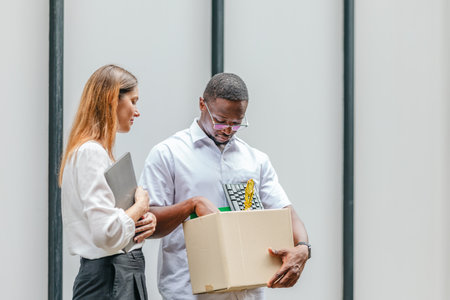 African male employee carries box of office belongings while female coworker encourages him...