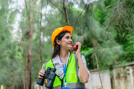 Female environmental surveyor observes forest conditions using binoculars, takes notes, communicates data. Environmental monitoring, biodiversity tracking, role of women in field forest assessment.の写真素材