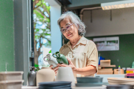 A senior Asian woman, the business owner, conducts quality control by visually inspecting ceramic products in a warehouse setting, emphasizing careful QC for retail and wholesale operations.の写真素材