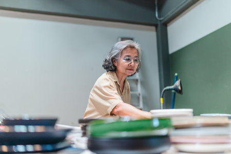 A senior Asian woman, the business owner, conducts quality control by visually inspecting ceramic products in a warehouse setting, emphasizing careful QC for retail and wholesale operations.の写真素材