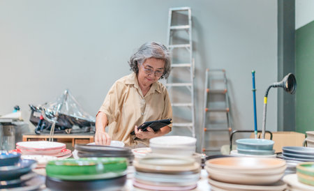 A senior Asian woman, the business owner, conducts quality control by visually inspecting ceramic...