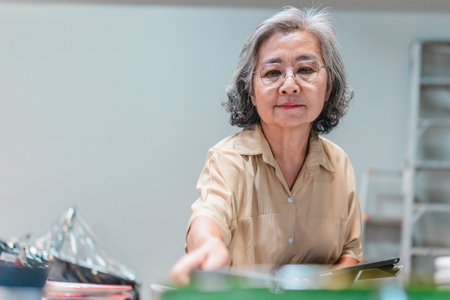 A senior Asian woman, the business owner, conducts quality control by visually inspecting ceramic products in a warehouse setting, emphasizing careful QC for retail and wholesale operations.の写真素材