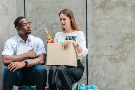 Female employee carries box of office belongings while African male coworker encourages her outdoors. Reflecting job loss during an economic downturn and power of workplace support and resilienceの写真素材