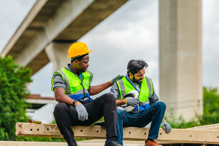 Two railway workers in safety vests and helmets take a break on concrete beams, showing fatigue while offering comfort and encouragement to one another on site.の写真素材