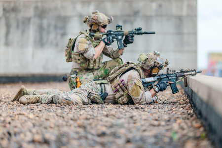 Tactical operators in camouflage uniforms take a prone and kneeling firing position with rifles behind a concrete barrier on a building rooftop during an urban training operation.