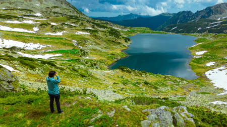 Landscape photography in an mountain range with a female photographer capturing the beautiful landscape with a glacial lake and mountains. Photographer capturing the beautiful sceneryの写真素材