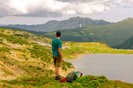 Photography of a male hiker in the mountains admiring a beautiful lake down in the valley in summer time with cloudy weather.の写真素材