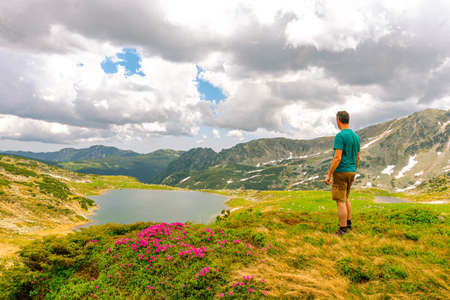 Photography of a male hiker in the mountains admiring a beautiful lake down in the valley in summer time with cloudy weather.の写真素材