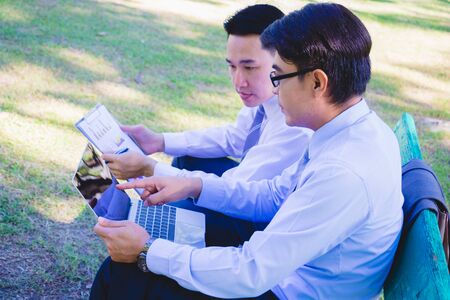 Businessman,They are sitting on bench in park.He is play notebook and  search internet.Another one holding a cup of coffee and talking about business in  relax time.Photo concept  business.の写真素材