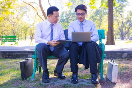 Businessman,They are sitting on bench in park.He is play notebook and  search internet.Another one holding a cup of coffee and talking about business in  relax time.Photo concept  business.の写真素材