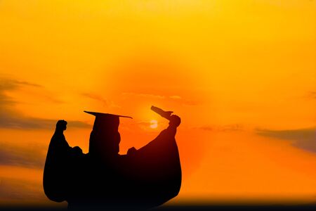 Silhouette photo.He s wearing  graduate's dress.He is standing and show hand.He is celebration education Graduation.He is happy in good time.の写真素材