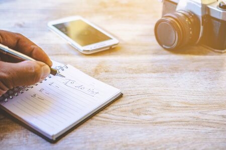 Hand holding a pen and notebook. Men are writing recorded on notebook a daily basis. Camera Phones and Accessories on old wood.の写真素材