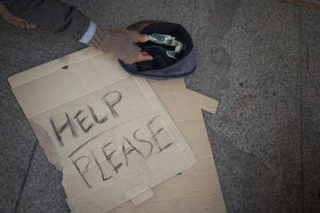 Homeless man is sitting down on walkway in town.He is receive dollar in hat with label âHelp pleaseâ word. He is hoping to have the better life.poverty,despair,Photo Sympathetic and hope concept.の写真素材