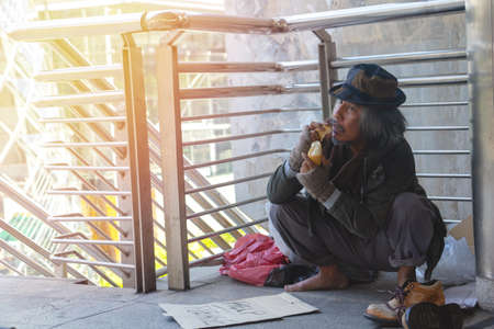 Homeless man is sitting down on walkway in town.He is raise hand  and eating bread.He is very...