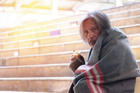 Homeless man is sitting down on staircase and walkway in town. He is eating bread. He is very unhappy. poverty, despair, Photo Sympathetic and hope concept.の写真素材