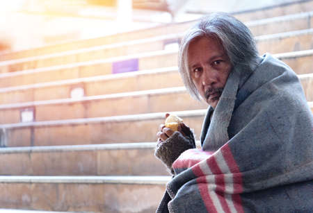 Homeless man is sitting down on staircase and walkway in town. He is eating bread. He is very unhappy. poverty, despair, Photo Sympathetic and hope concept.の写真素材