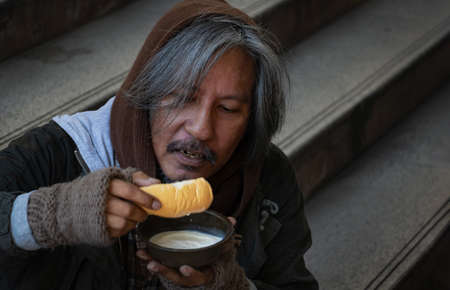 Homeless man is sitting down on staircase in town.He is eat bread and milk.He is very hungry and unhappy.poverty,despair, Photo Sympathetic and hope concept.の写真素材
