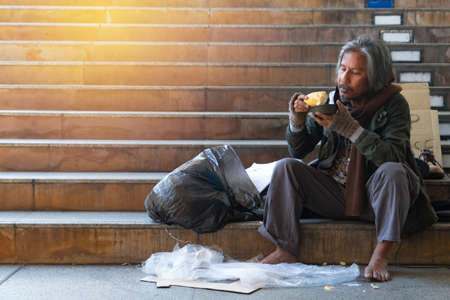 Homeless man is sitting down on staircase in town.He is eat bread and milk.He is very hungry and unhappy.poverty,despair, Photo Sympathetic and hope concept.の写真素材