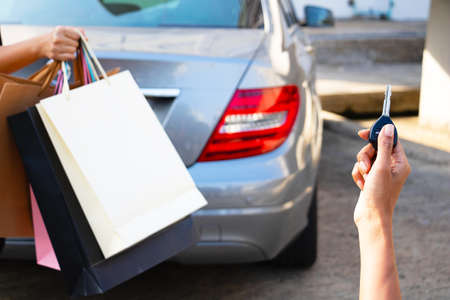 The hand woman holding a car key and back of a woman carrying a shopping bag with the car in the background. They returned from shopping.buy,sell,Photo concept Lifestyle and Shopping.の写真素材