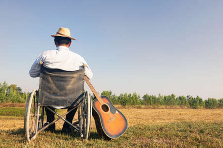 Young disabled man and guitar with field background.He is raising hand looking into sky.He is wearing a hat and sitting on wheelchair. despair,lonely,hope.Photo concept depression and Patient.の写真素材