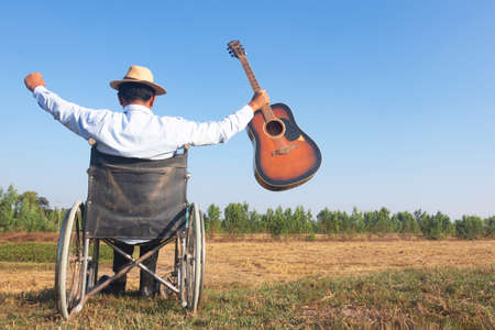 Young disabled man and guitar with field background.He is raising hand looking into sky.He is wearing a hat and sitting on wheelchair. despair,lonely,hope.Photo concept depression and Patient.の写真素材